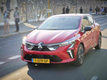 Een rode Mitsubishi Colt met een geel kenteken rijdt door een stadsstraat met een met muurschildering bedekte muur op de achtergrond.