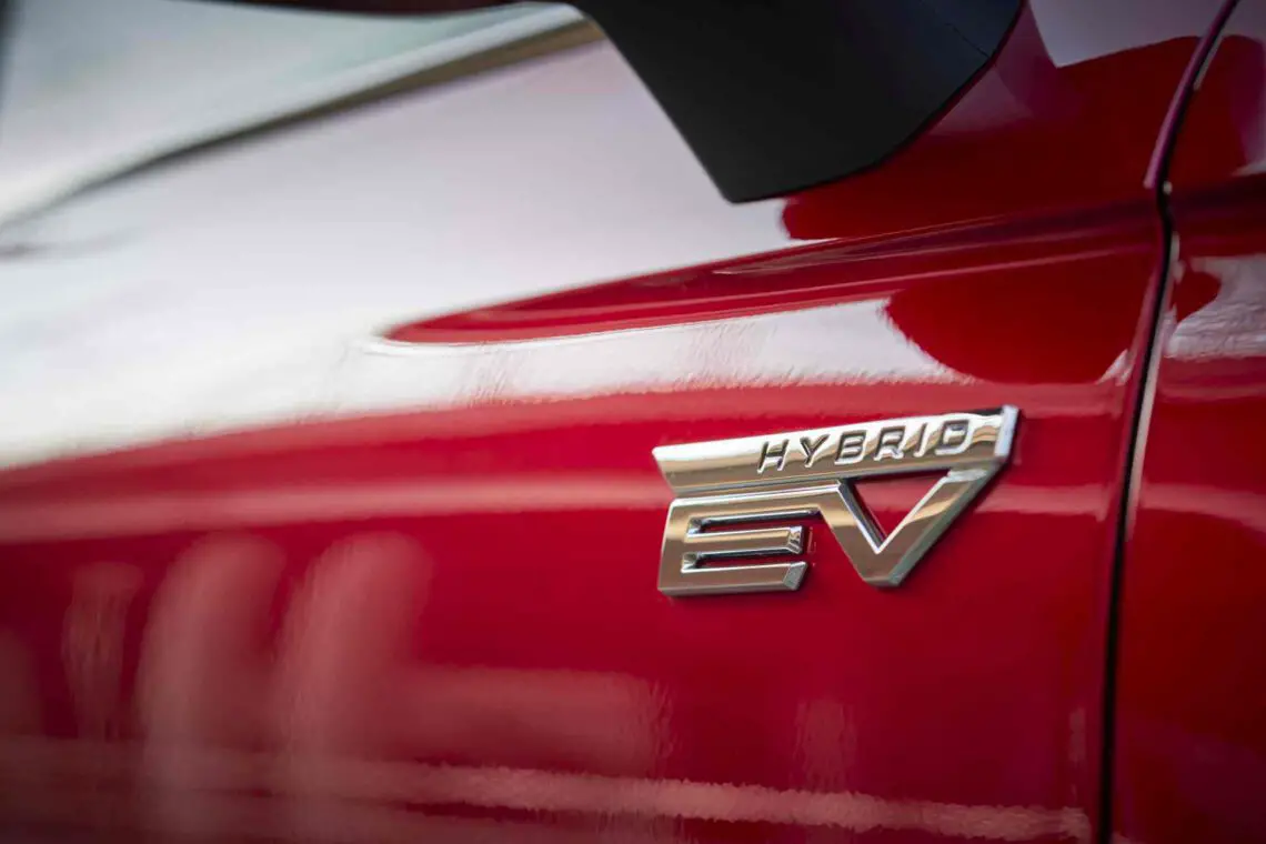 Close-up van het 'HYBRID EV'-embleem op de zijkant van een rode Mitsubishi Colt, wat aangeeft dat het een hybride elektrisch voertuig is.