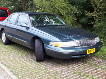 Een donkergroene Lincoln Continental sedan met een gele kentekenplaat staat geparkeerd op een berm met gras en kasseien. Daarachter is gedeeltelijk een rood voertuig zichtbaar. Bomen en struiken zijn op de achtergrond.