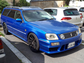 Op een parkeerplaats tussen andere auto's staat een blauwe Nissan Stagea met een aangepaste voorbumper.