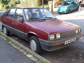 Een rode Renault 11 vierdeurs sedan staat geparkeerd in een straat in een buitenwijk. Op de achtergrond is een blauw busje zichtbaar. Op het kenteken van de sedan staat "418 BZX 95.