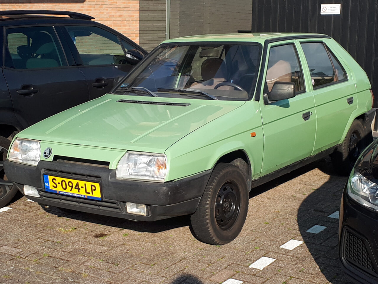 Een groene Skoda Favorit vijfdeurs hatchback staat geparkeerd op een parkeerplaats tussen twee andere voertuigen. Er staat een kentekenplaat met de tekst "S-094-LP".