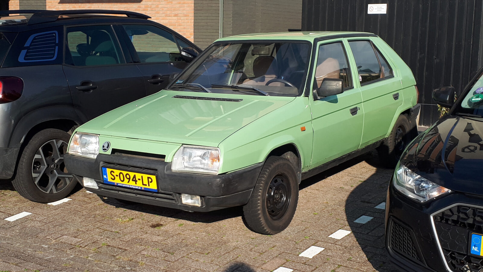 Een groene Skoda Favorit vijfdeurs hatchback staat geparkeerd op een parkeerplaats tussen twee andere voertuigen. Er staat een kentekenplaat met de tekst "S-094-LP".