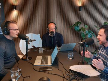 Drie mannen met koptelefoon zitten rond een tafel met microfoons en laptops en zijn bezig met het opnemen van een aflevering van de AutoRAI Podcast in een studio met houten lambrisering. De discussie, onder leiding van Fons Nijkrake, gaat dieper in op BYD in Nederland.