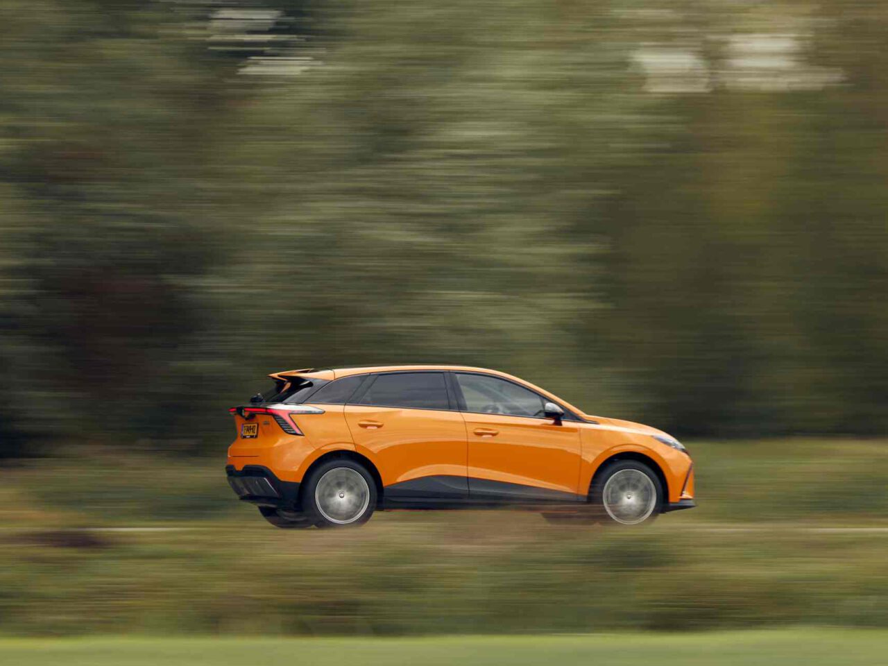 Een MG4 Electric XPOWER SUV in een levendige oranje tint rijdt met hoge snelheid door een groene en onscherpe achtergrond.