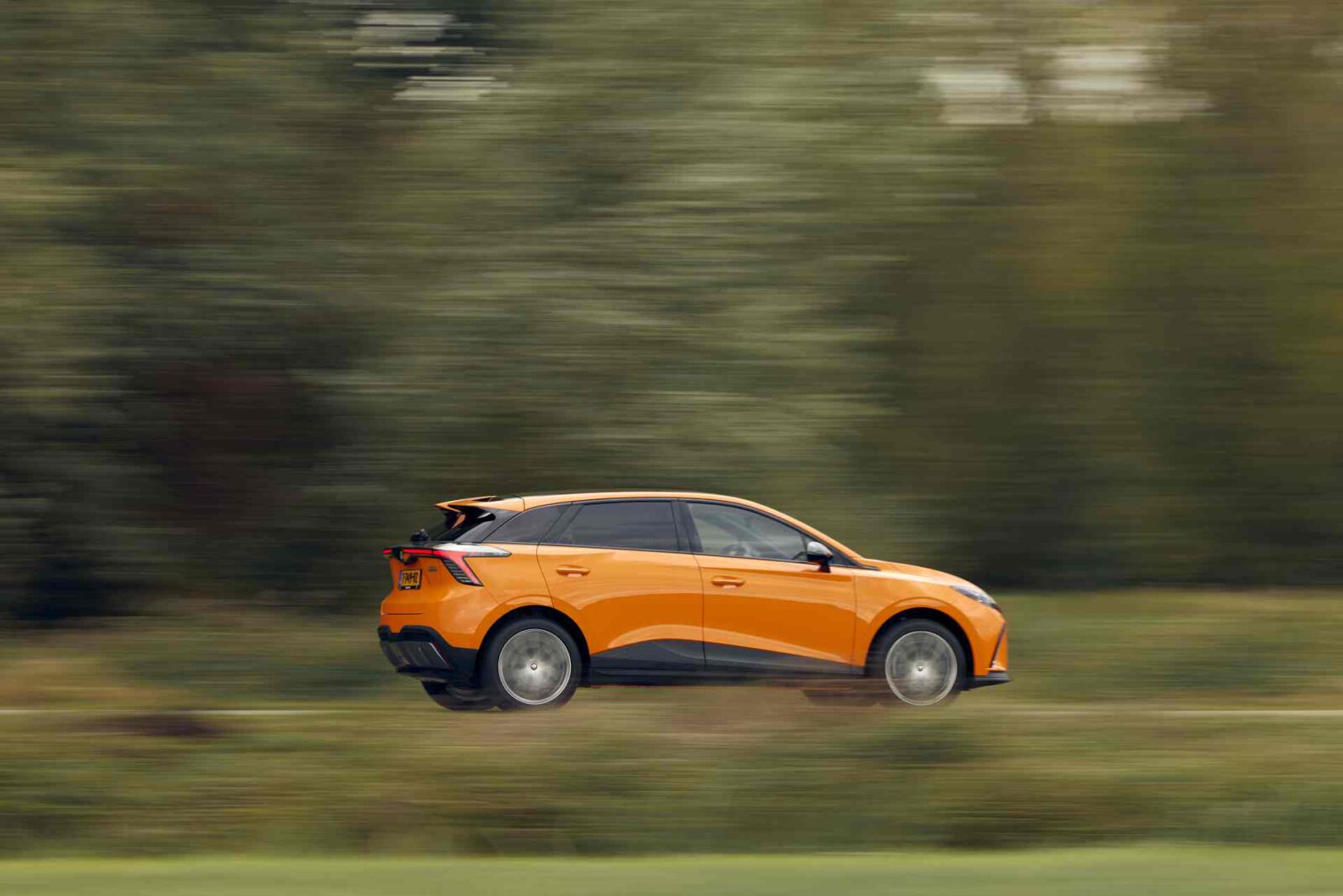 Een MG4 Electric XPOWER SUV in een levendige oranje tint rijdt met hoge snelheid door een groene en onscherpe achtergrond.