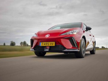 Een rode MG4 Electric XPOWER-auto met Nederlands kenteken "T-495-VV" die op een open weg rijdt, tegen een achtergrond van een bewolkte lucht en een groen groen landschap.