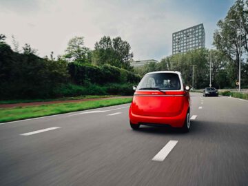 Een strakke rode Microlino rijdt over een meerbaansweg omringd door groen en moderne gebouwen.