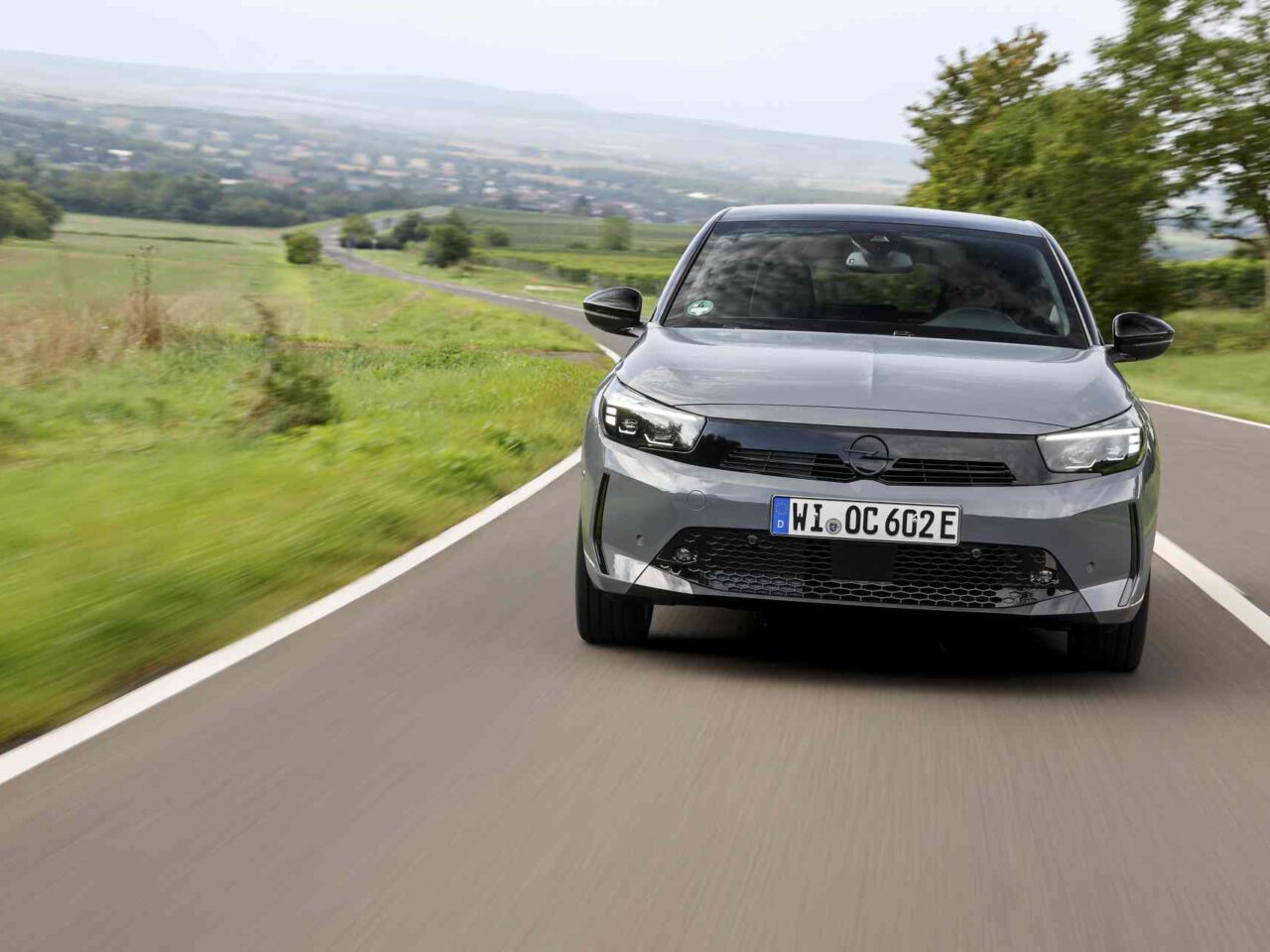 Een grijze Opel Corsa rijdt op een landelijke weg met twee rijstroken, met groene velden en bomen aan de zijkanten. Op het kenteken aan de voorzijde van de auto staat "W1 OC 622E.