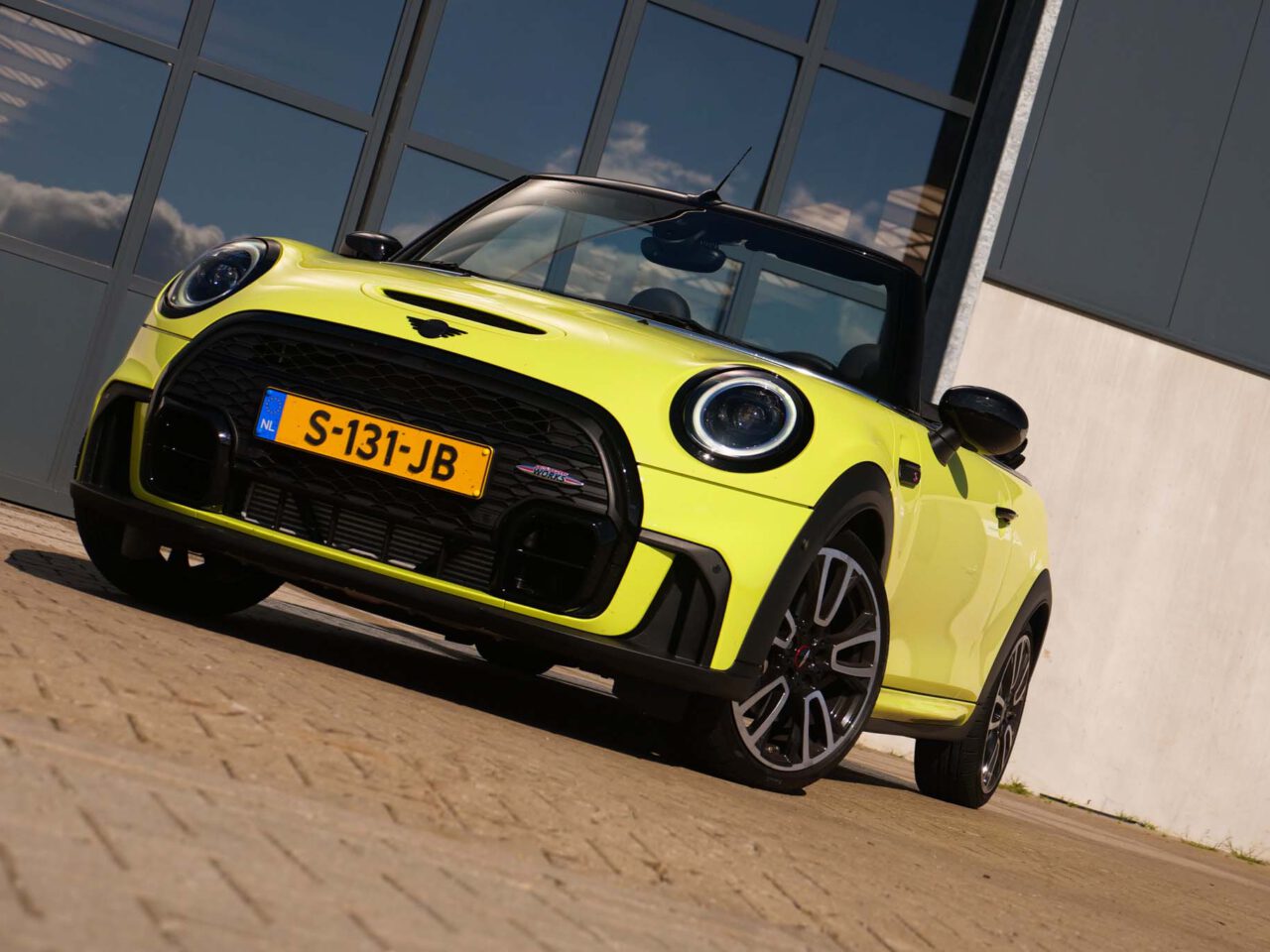 Een limoengroene Mini Cooper-cabriolet staat geparkeerd op een verhard terrein voor een modern gebouw met grote ramen. Op het kenteken van de auto staat "S-131-JB.