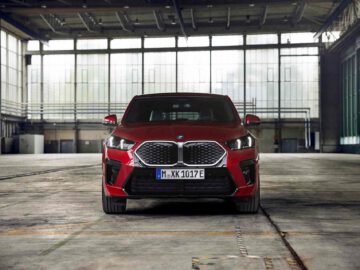 Vooraanzicht van een rode BMW X2 geparkeerd in een ruim industrieel pakhuis met grote ramen op de achtergrond.