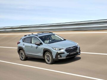 Een grijze Subaru Crosstrek rijdt op een meerbaansweg met metalen leuningen, onder een heldere hemel.