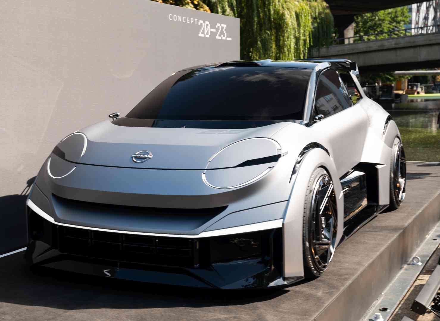 Futuristische zilveren auto buiten tentoongesteld met een bordje op de zijkant met de tekst "Nissan Concept 20-23". De auto heeft een strak, modern ontwerp en grote, ronde koplampen.