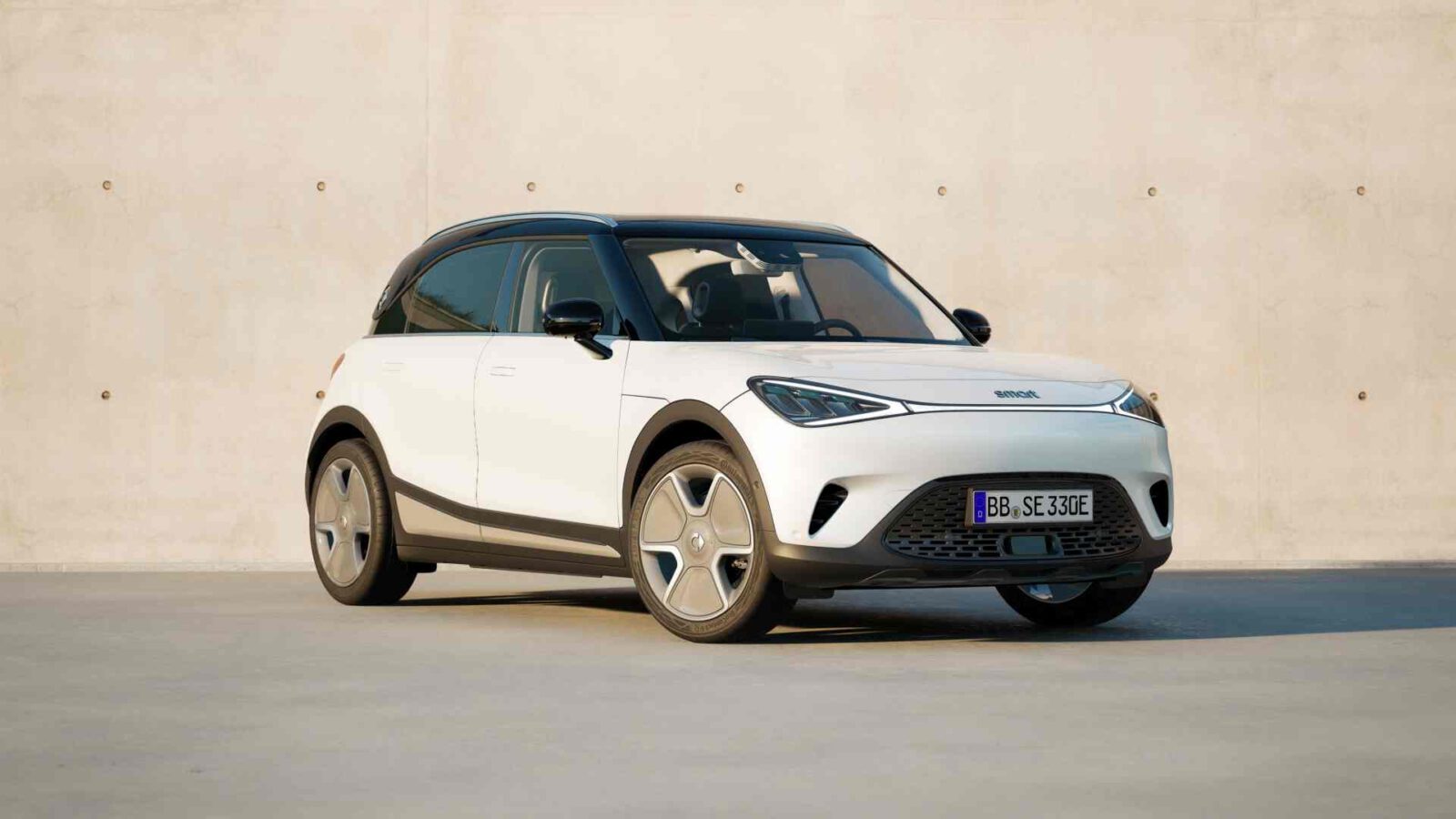 Een strakke, witte elektrische compacte SUV met vijfspaaks wielen en donker getinte ramen staat netjes geparkeerd voor een beige betonnen muur.