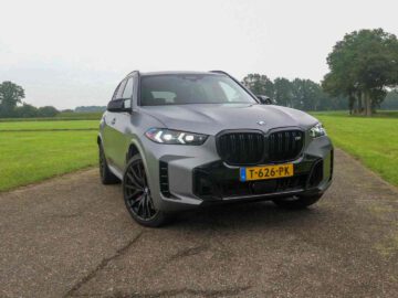 Een grijze BMW X5 SUV met Nederlands kenteken "T-626-PK" staat geparkeerd op een verhard pad in een landelijk gebied, waarbij gras en bomen de perfecte achtergrond vormen voor dit prachtige voertuig.