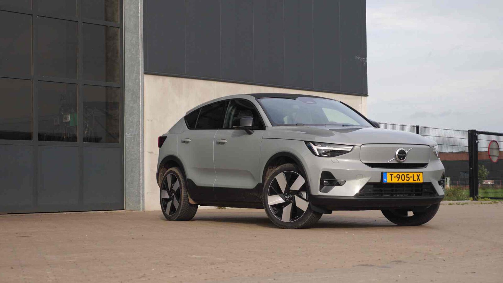 Een zilverkleurige Volvo C40 SUV staat geparkeerd op een verhard oppervlak nabij een groot gebouw met ramen. Het voertuig heeft een geel kenteken met het nummer "T-905-LX.