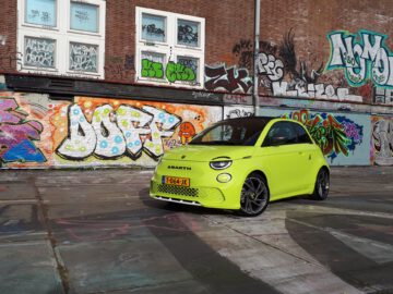 Een limoengroene Abarth 500e-auto staat geparkeerd voor een met graffiti bedekte muur op een met graffiti bedekte stoep. Op het kenteken van de auto staat "T-064-JJK.