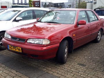 Een geparkeerde rode Hyundai Sonata met een geel Europees kenteken staat tussen witte auto's op een buitenparkeerplaats.