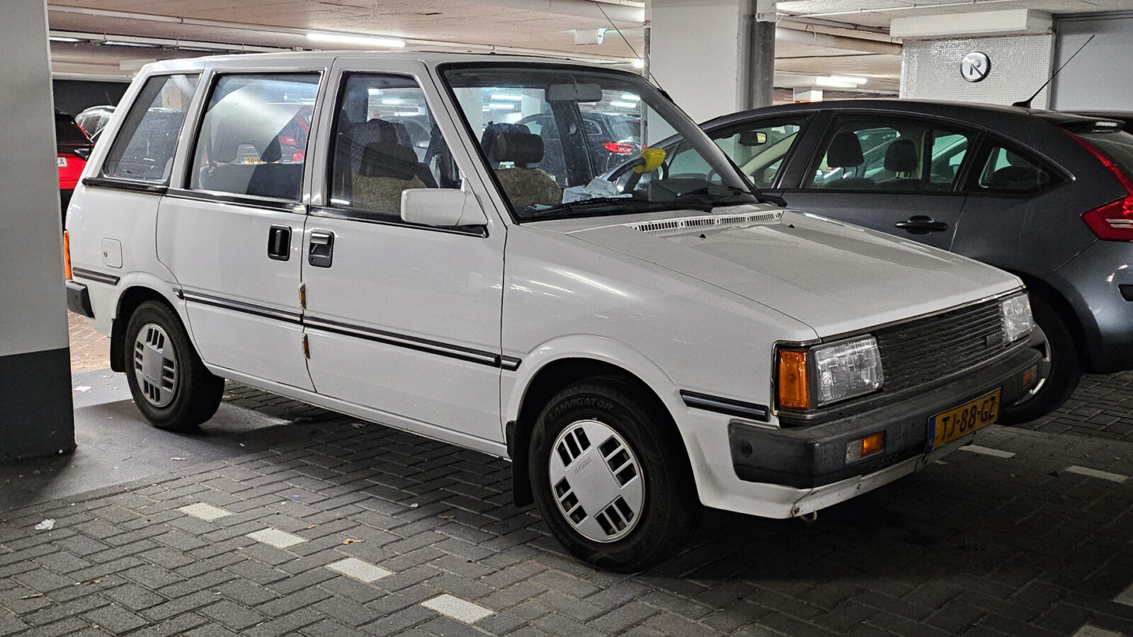 Een witte vintage Nissan Prairie staat geparkeerd in een overdekte parkeergarage naast andere voertuigen. De auto heeft een boxy design met rechthoekige koplampen en een Nederlands kenteken.