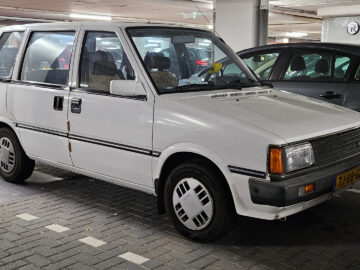 Een witte vintage Nissan Prairie staat geparkeerd in een overdekte parkeergarage naast andere voertuigen. De auto heeft een boxy design met rechthoekige koplampen en een Nederlands kenteken.