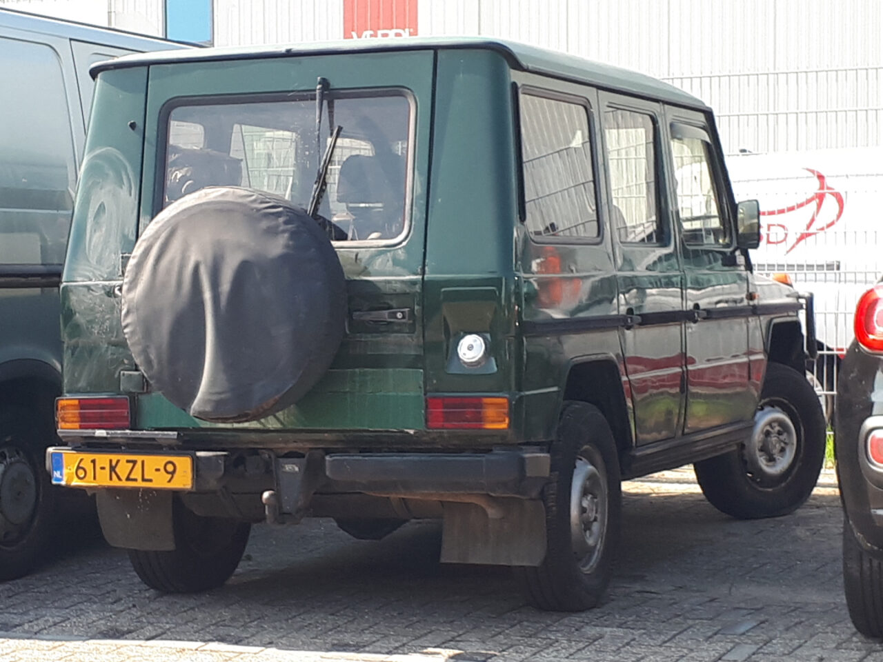 Een groene Mercedes-Benz G-Klasse SUV staat geparkeerd op een parkeerplaats naast een grijs voertuig en een zwarte auto. De G-Klasse heeft een afgedekt reservewiel achterop en een geel kenteken met nummer 61-KZL-9.