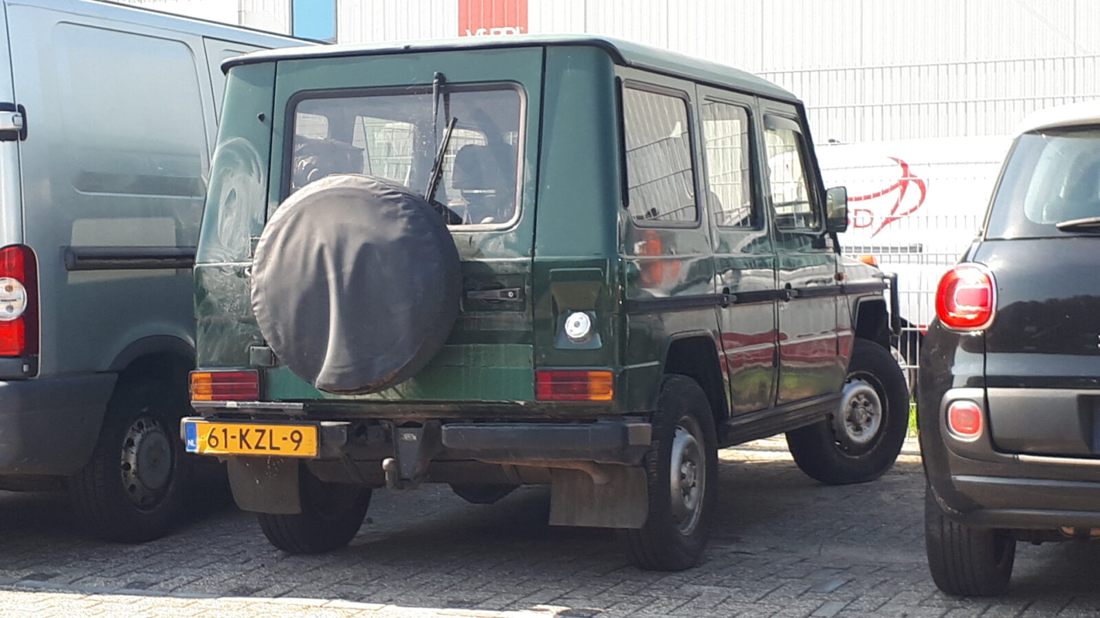 Een groene Mercedes-Benz G-Klasse SUV staat geparkeerd op een parkeerplaats naast een grijs voertuig en een zwarte auto. De G-Klasse heeft een afgedekt reservewiel achterop en een geel kenteken met nummer 61-KZL-9.