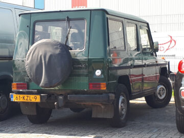 Een groene Mercedes-Benz G-Klasse SUV staat geparkeerd op een parkeerplaats naast een grijs voertuig en een zwarte auto. De G-Klasse heeft een afgedekt reservewiel achterop en een geel kenteken met nummer 61-KZL-9.