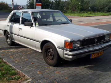 Een geparkeerde zilverkleurige Toyota Crown sedan met een geel kenteken wordt vastgelegd op een verhard terrein naast een rijbaan. Het voertuig lijkt een ouder model te zijn.