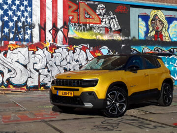 Een gele Jeep Avenger SUV staat geparkeerd voor een met graffiti bedekte muur. De graffiti bevat een Amerikaanse vlag, verschillende tags en illustraties. Het voertuig beschikt over een kenteken "T-538-FH.