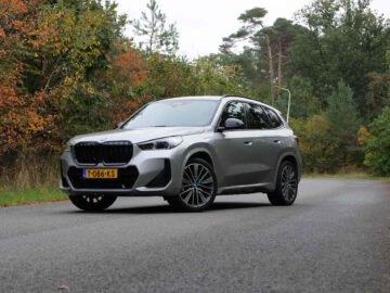 Een BMW X1 met geel kenteken staat geparkeerd op een verharde weg omgeven door bomen en groen.