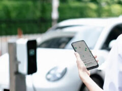 Persoon die een smartphone vasthoudt met de GRID-app terwijl hij naast een witte elektrische auto staat, beladen met boodschappen, die aan het opladen is bij een station.