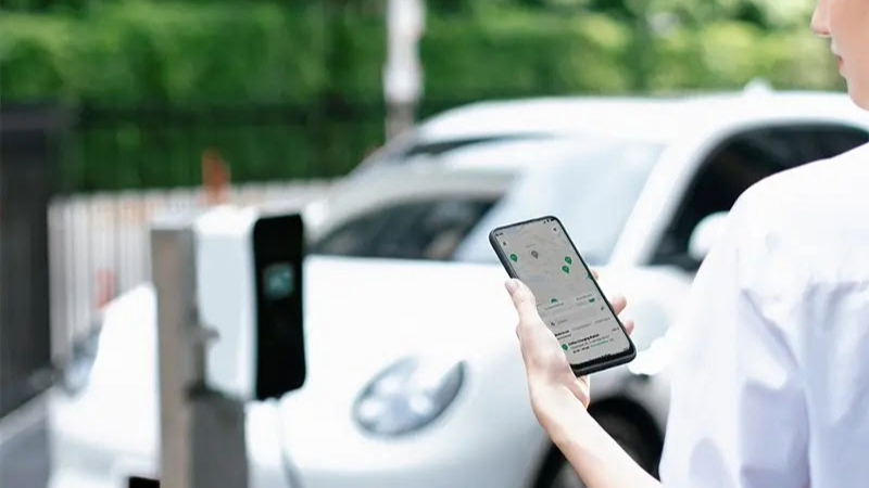 Persoon die een smartphone vasthoudt met de GRID-app terwijl hij naast een witte elektrische auto staat, beladen met boodschappen, die aan het opladen is bij een station.