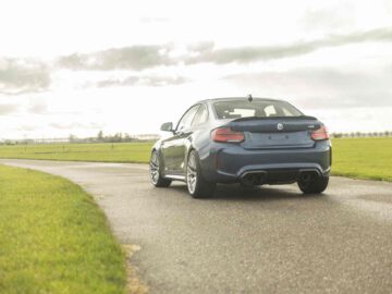Een donkerblauwe BMW X2 rijdt weg op een smalle asfaltweg omgeven door groene velden onder een bewolkte hemel.