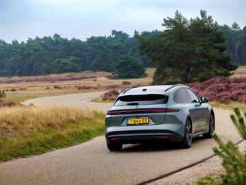 Een zilveren NIO ET5 met een geel kenteken rijdt over een smalle weg in een schilderachtig landelijk gebied, omlijst door bomen en paarse heide op de achtergrond.