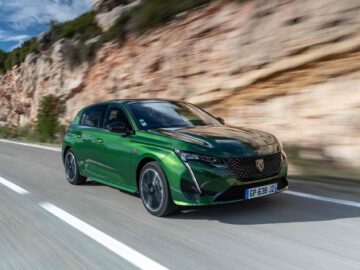 Een Peugeot e-308 rijdt op een weg naast een rotsachtige klif onder een blauwe lucht met wolken.