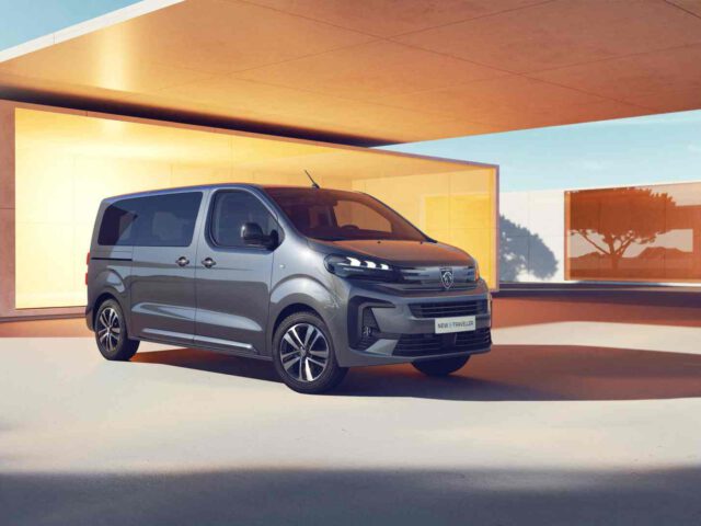 Een grijze Peugeot e-Traveller staat geparkeerd voor een modern gebouw onder een pergola, waarbij het zonlicht de buitenkant benadrukt.