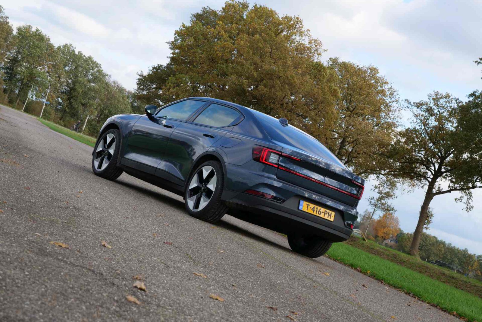 Een donkergrijze elektrische Polestar-sedan geparkeerd op een verhard oppervlak, omgeven door bomen en groen op een bewolkte dag.