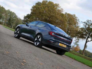 Een donkergrijze elektrische Polestar-sedan geparkeerd op een verhard oppervlak, omgeven door bomen en groen op een bewolkte dag.