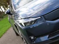 Close-up van het voorste gedeelte van een blauwe Polestar sedan geparkeerd aan de kant van de weg, waarbij de koplamp, de grille en een deel van het voorspatbord benadrukt worden. Op de achtergrond zijn gras en bomen zichtbaar.