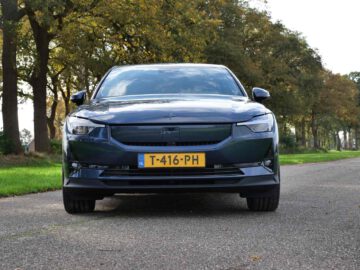 Vooraanzicht van een donkergekleurde elektrische Polestar 2-auto met zichtbaar kenteken "T-416-PH", geparkeerd op een met bomen omzoomde weg onder een bewolkte hemel.