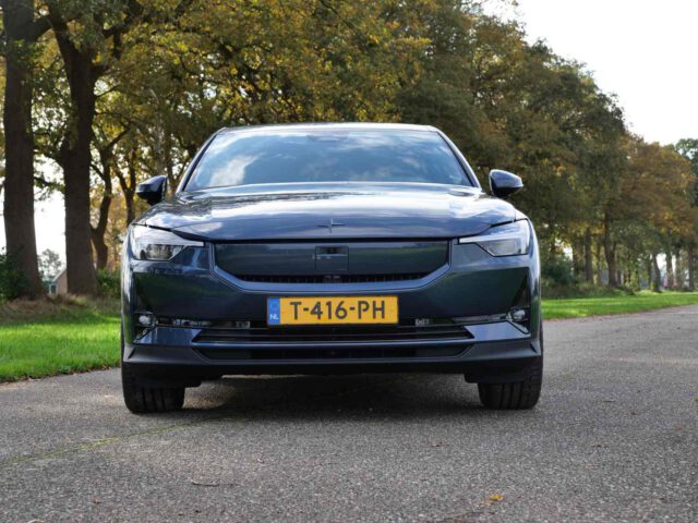 Vooraanzicht van een donkergekleurde elektrische Polestar 2-auto met zichtbaar kenteken "T-416-PH", geparkeerd op een met bomen omzoomde weg onder een bewolkte hemel.