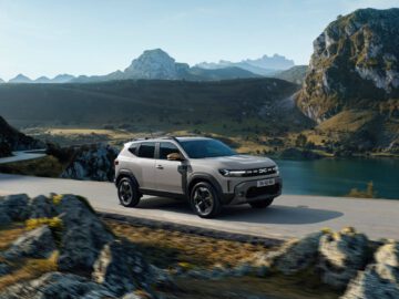 Een Dacia Duster 2024 rijdt over een bochtige weg met rotswanden en een meer op de achtergrond onder een heldere hemel.