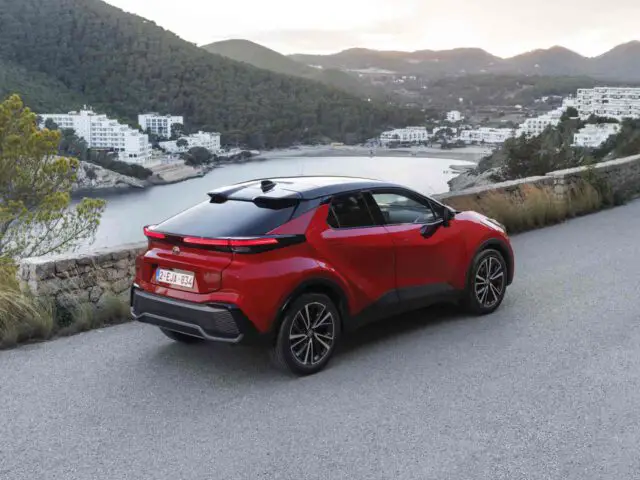 Een rode Toyota C-HR, een compacte crossover, geparkeerd op een schilderachtige weg met uitzicht op een kustlandschap met heuvels en gebouwen op de achtergrond.