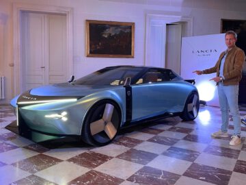 Een persoon staat naast een strakke, futuristische Lancia Pu+Ra HPE in een goed verlichte kamer met een geruite vloer. De auto heeft een modern design met scherpe koplampen.