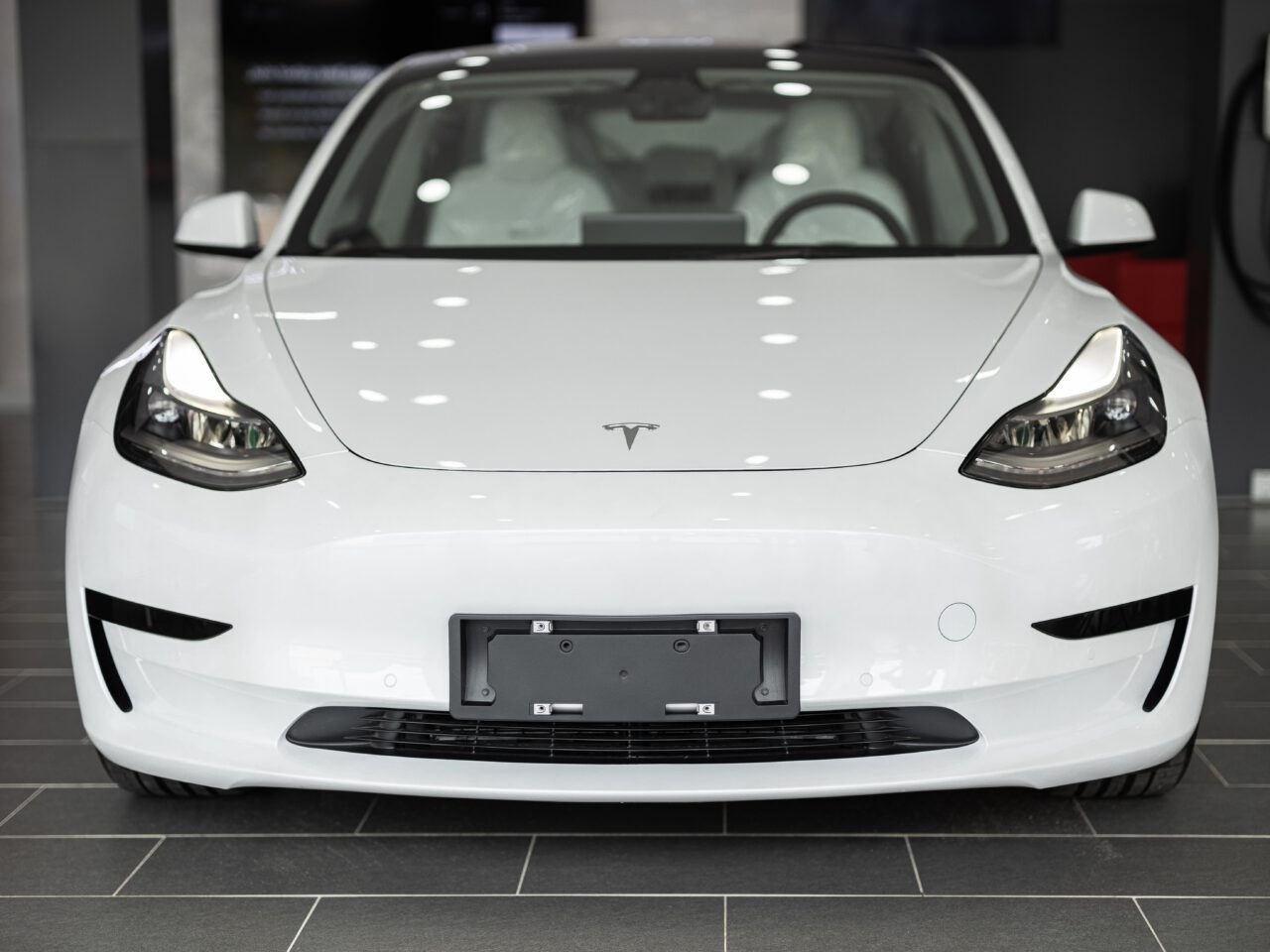 Van voren wordt een witte Tesla elektrische auto getoond, binnen geparkeerd met een laadstation zichtbaar op de achtergrond.