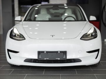 Van voren wordt een witte Tesla elektrische auto getoond, binnen geparkeerd met een laadstation zichtbaar op de achtergrond.