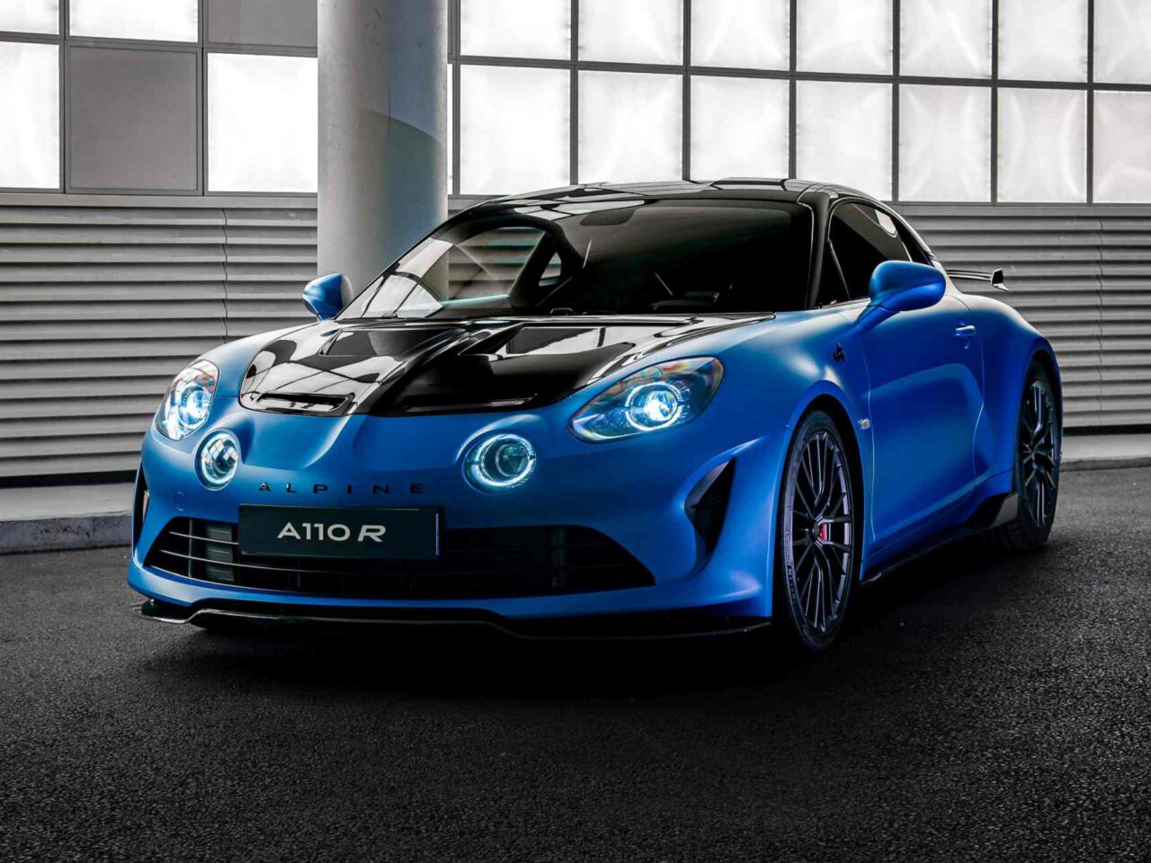 Een strakke blauwe Alpine A110 R-sportwagen staat binnen geparkeerd onder een dak met rastervensters en laat zijn indrukwekkende aerodynamica en opvallende LED-koplampen zien.