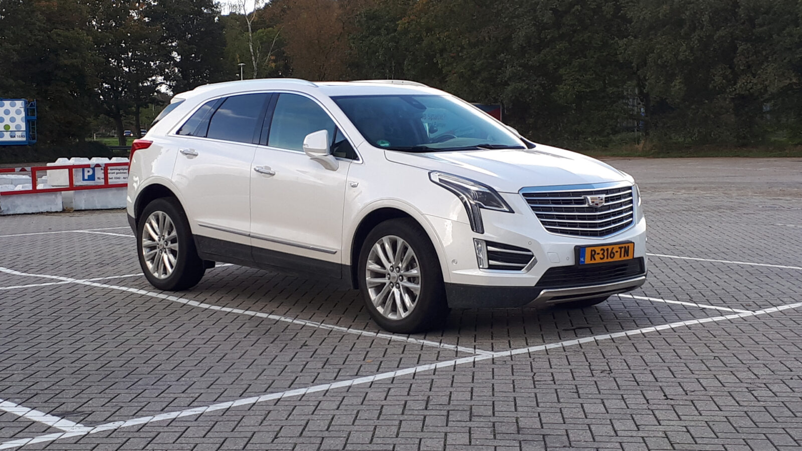 Een witte Cadillac XT5 SUV staat geparkeerd op een vrijwel leeg terrein met bomen op de achtergrond. Het voertuig heeft Nederlands kenteken.
