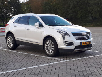 Een witte Cadillac XT5 SUV staat geparkeerd op een vrijwel leeg terrein met bomen op de achtergrond. Het voertuig heeft Nederlands kenteken.