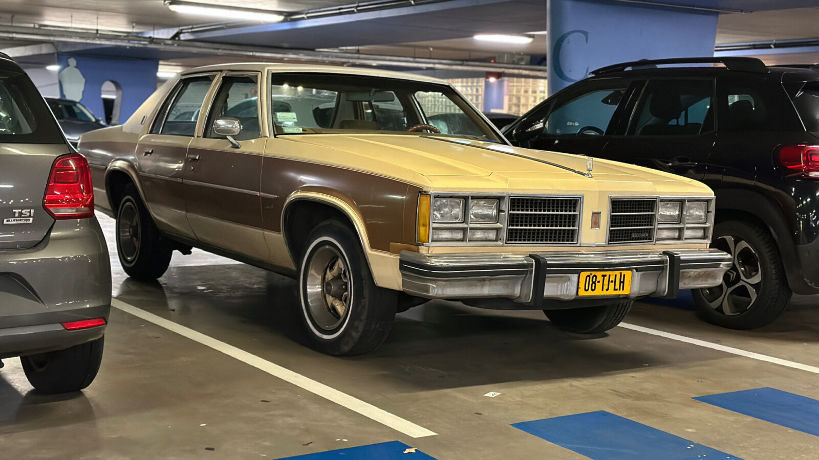 Een beige en bruine vintage Oldsmobile Delta 88 staat geparkeerd op een overdekte parkeerplaats, geflankeerd door een zilveren auto aan de linkerkant en een zwarte SUV aan de rechterkant. De auto heeft een geel kenteken met de tekst "08-TL-H".